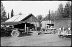 Vägstation AC19, Storuman. Förrådsbyggnader. Fronten av en väghyvel skymtar till vänster, fronten på personbil till höger. Vägsladd i mitten. Upplag av diverse materiel.