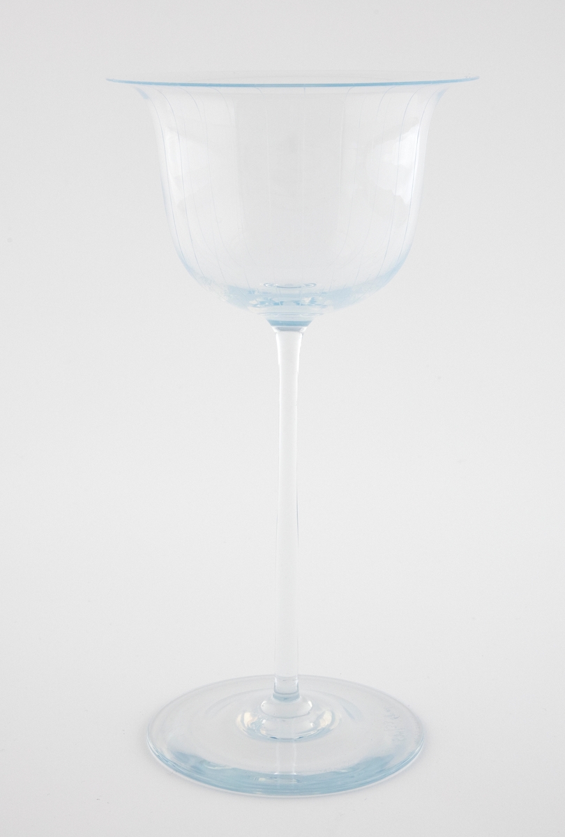 Alterkalk i gjennomskinnelig glass med opaliserende partier. Klokkeformet kupa med bred munning, som hviler på en høy og smal stett og sirkulær fotskive. Kupa er dekorert graverte vertikale linjer.