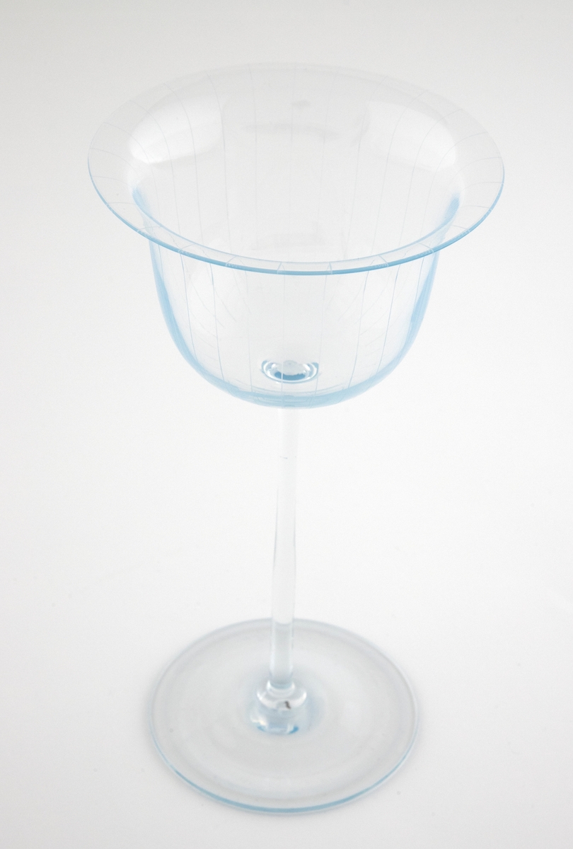 Alterkalk i gjennomskinnelig glass med opaliserende partier. Klokkeformet kupa med bred munning, som hviler på en høy og smal stett og sirkulær fotskive. Kupa er dekorert graverte vertikale linjer.