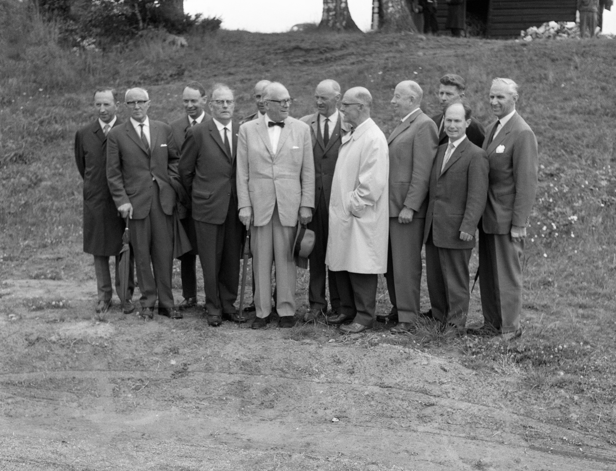 Bilde tatt i forbindelse med åpningen av Klokkerfossbrua i 1964. Brua over Klokkerfossen i Glomma, Elverum, Hedmark.
Norsk Skogmuseums styre, fotografert ved vegen vest for klokkergarden Fossum i Elverum i samband med representantskapsmøtet som ble avviklet 30. juni 1964. Forsamlinga besto av tolv dress- og til dels frakkekledde menn. Fra venstre ser vi direktør Ivar Aavatsmark fra Norges Skogeierforbund, advokat Finn Krog, direktør Per Krog, forretningsfører Klaus Kjelsrud (styrets nestleder), direktør Kaare Sparby, diplomingeniør Hans Th. Kiær (statens representant i styret, styreleder), statskogsjef Eyvind Wisth (representantskapets leder), advokat Finn Krog, disponent Oskar Moripen, generalsekretær Magne Midttun, konservator Tore Fossum og direktør Wilhelm Matheson (styresekretær). På haugen i bakgrunnen ser vi en del andre menn, sannsynligvis stort sett medlemmer av museets representantskap. Vi ser også gavlen på en laftet bygning som var rødmalt, men hadde kvite endeflater på novene og kvite vindskier. Dette er sannsynligvis det lille fjøset som seinere ble flyttet til den søndre delen av tunet på klokkergarden Fossum, der den ble tatt i bruk som vedskåle. At det ble bestilt fotograf til å ta et gruppebilde av museumsstyret nettopp i 1964 hadde sannsynligvis sammenheng med at museets styrende organer den nevnte dagen markerte Skogbruksmuseets tiårsjubileum. Styre- og representantskapsmøtene ble avviklet på hotell Central, hvor deltakerne blant annet fikk se de nye kulturhistoriske filmene museet hadde fått produsert om lågåsildfisket nederst i Gudbrandsdalslågen og filmen «Liv og husvær i skogen», som handlet om skogsarbeidernes kår. Deretter var det omvisning på museumsområdet, hvor det var etablert utstillinger i klokkergardslåven og bygd ny bru over Klokkerfossen til Prestøya. Det var sannsynligvis under denne delen av arrangementet dette fotografiet ble tatt. Jubileumsmarkeringa med styre og representantskap ble avsluttet med middag i Stemsrudstua på Glomdalsmuseet, der fruene Ragnhild Fossum og Ester Krog var vertinner. Her holdt flere av de avbildete, blant annet styreleder Hans Th. Kiær, taler som oppsummerte hva som var utrettet gjennom de første ti åra og antydet utviklingsplaner, blant annet for et nytt museumsbygg.