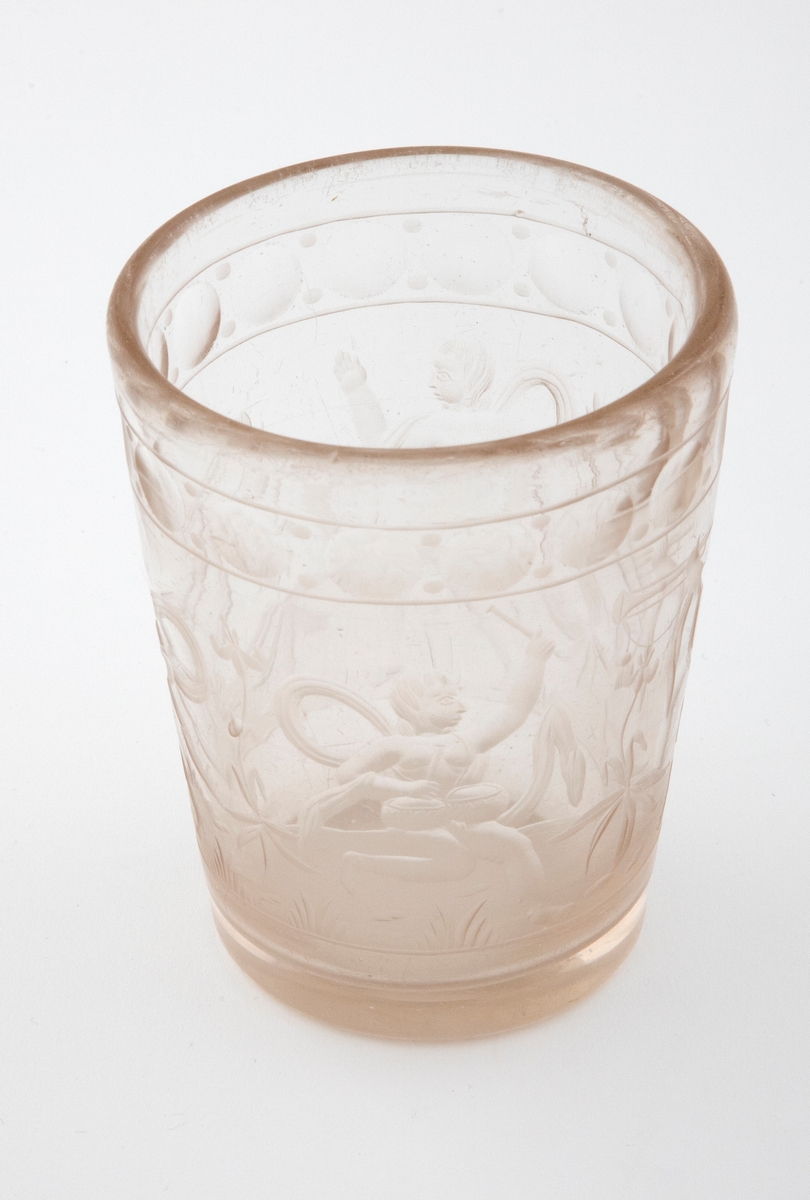 Tilnærmet sylindrisk beger i tykt klart glass med svakt koniske vegger. Yttersiden er dekorert med sliping og gravering. Øverst er det fremstilt en frise med sirkler. Deretter følger fremstillinger av fire nakne barneskikkelser - omgitt av flagrende drapperier og planter - som spiller på ulike musikkinstrumenter.