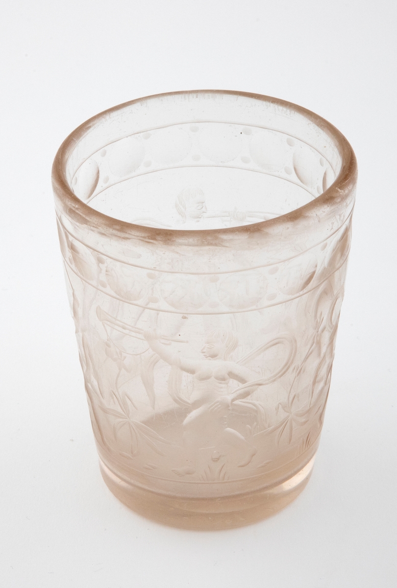Tilnærmet sylindrisk beger i tykt klart glass med svakt koniske vegger. Yttersiden er dekorert med sliping og gravering. Øverst er det fremstilt en frise med sirkler. Deretter følger fremstillinger av fire nakne barneskikkelser - omgitt av flagrende drapperier og planter - som spiller på ulike musikkinstrumenter.