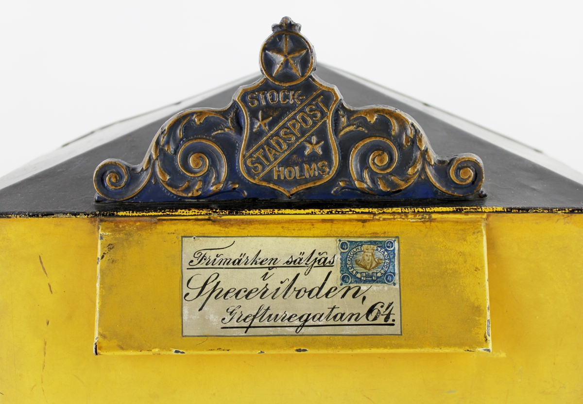 Brevlåda med sluttande, brutet tak. I främre delen av taket ett av Stockholms stadspost emblem. Brevlådans tak och ramen runt brevlådans botten är svart i övrigt är brevlådan gul. På inläggsklaffen ett vitt kuvert med ett av Stadspostens frimärken och texten "Frimärken säljas i Speceriboden Grefturegatan 64".
Där under en liten glasruta avsedd för visning av tömningstider. Brevlådans front har inskription. Brevlådan tömmes från botten med en tömningsväska. Ett lås är centrerat i lådans nederkant. Nyckeln har PM 1642.
