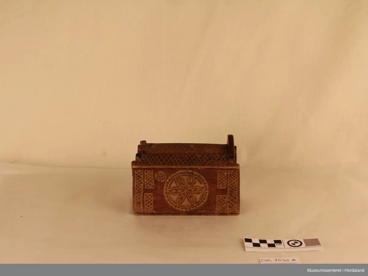 Liten geometrisk forma ekse med skyvelokk i toppen. Lokket er ovalt forma, med et "kryss" mønster, som går frå lokket og over på sidene. Det er ei skåra inn ei blomme på kvar av sidene på langs av esken. Ene kortsiden har ein avlang høg trekanta side, men langsiden er denne ødelagt. Esken er farga mørkebrun.