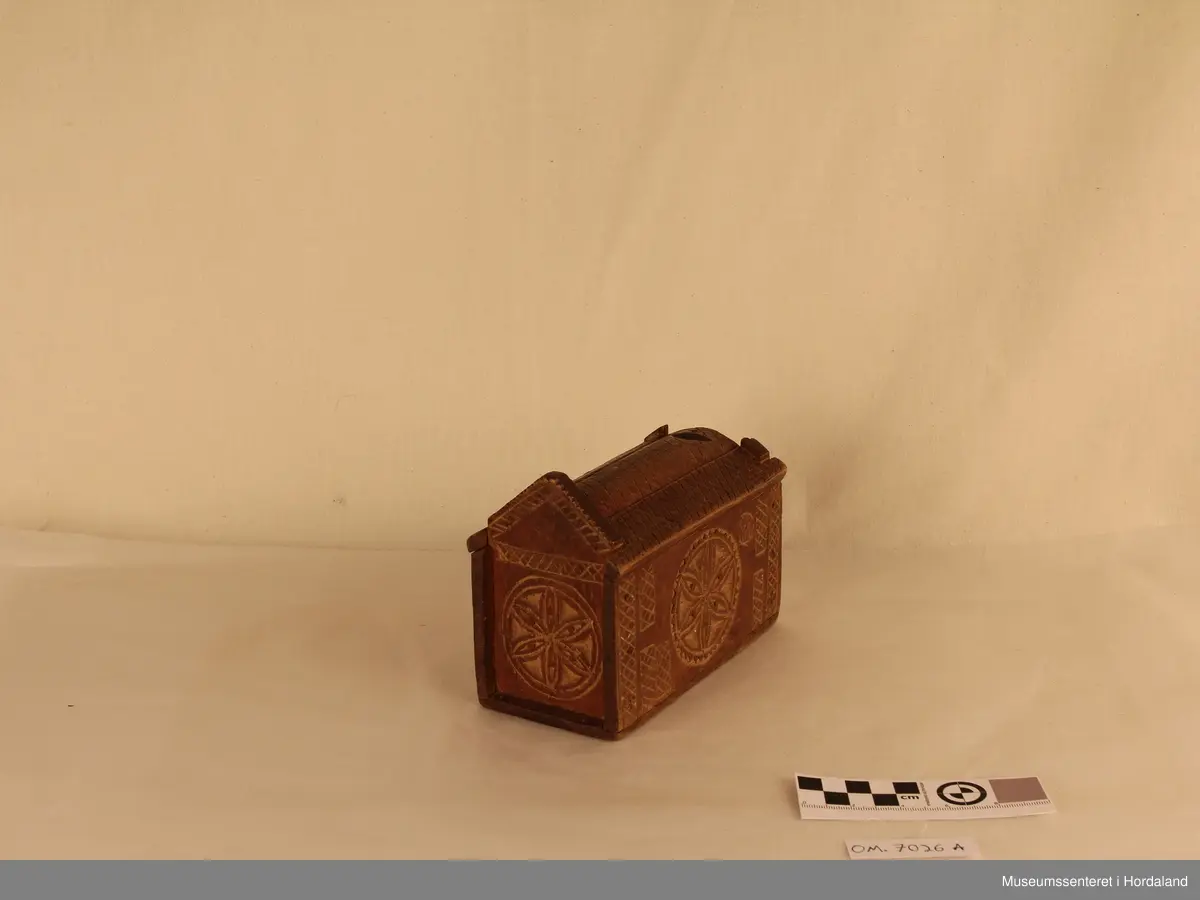 Liten geometrisk forma ekse med skyvelokk i toppen. Lokket er ovalt forma, med et "kryss" mønster, som går frå lokket og over på sidene. Det er ei skåra inn ei blomme på kvar av sidene på langs av esken. Ene kortsiden har ein avlang høg trekanta side, men langsiden er denne ødelagt. Esken er farga mørkebrun.