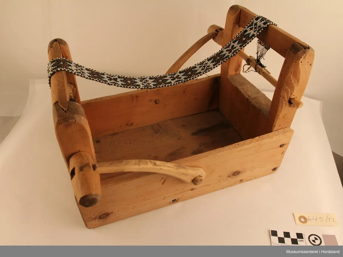 Perlevev i ulakkert tre. Veven er enkelt utført utan ornamentikk. Veven har et perlearbeid med åttebladroser i kvite og svarte perler. Bakgrunnen i motivet er i gult, grønt og blått. Langs kantane har bandet en rad med vekselvis kvite og svarte perler, innafor denne er det ein rad kvite perlar.