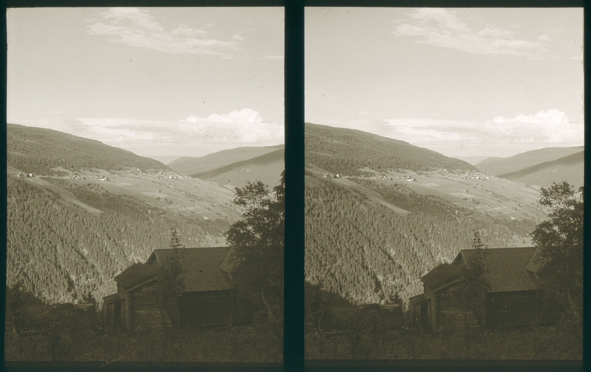 Utsikt over fjelllandskap med skog og dyrket mark. Et gammelt trebygg i forgrunnen. Tilhører Arkitekt Hans Grendahls samling av stereobilder.