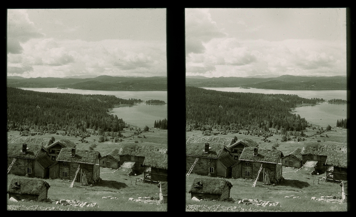 Gårdstun i Telemark. Det står grupper med laftede uthus på tunet. I bakgrunnen viser landskapet med innsjøen Totak. Tilhører Arkitekt Hans Grendahls samling av stereobilder.