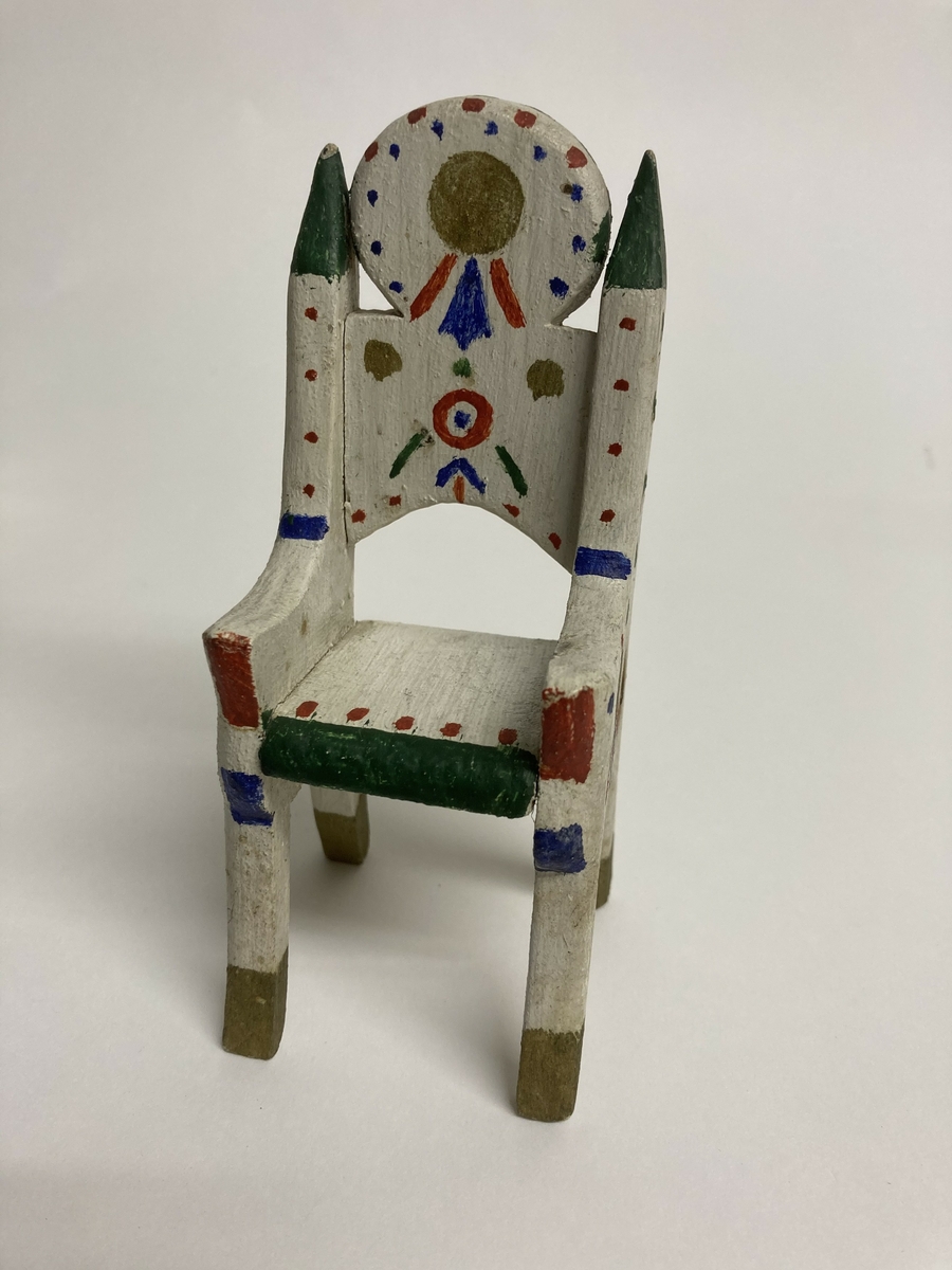 Stol til dokkemøblement i fem deler. Denne stolen er litt mindre enn de tre andre. Møblene er hvitmalte med dekor i ulike farger. 
Møblementet er fra givers tidlige barndom, muligens 1928-1930. Møblene var til hennes første dokkestue, bygget av far av en appelsinkasse. 