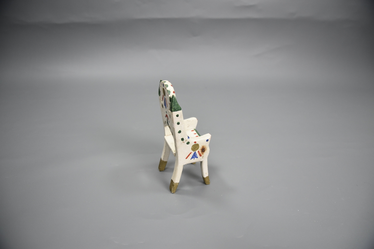 Stol til dokkemøblement i fem deler. Denne stolen er litt mindre enn de tre andre. Møblene er hvitmalte med dekor i ulike farger. 
Møblementet er fra givers tidlige barndom, muligens 1928-1930. Møblene var til hennes første dokkestue, bygget av far av en appelsinkasse. 