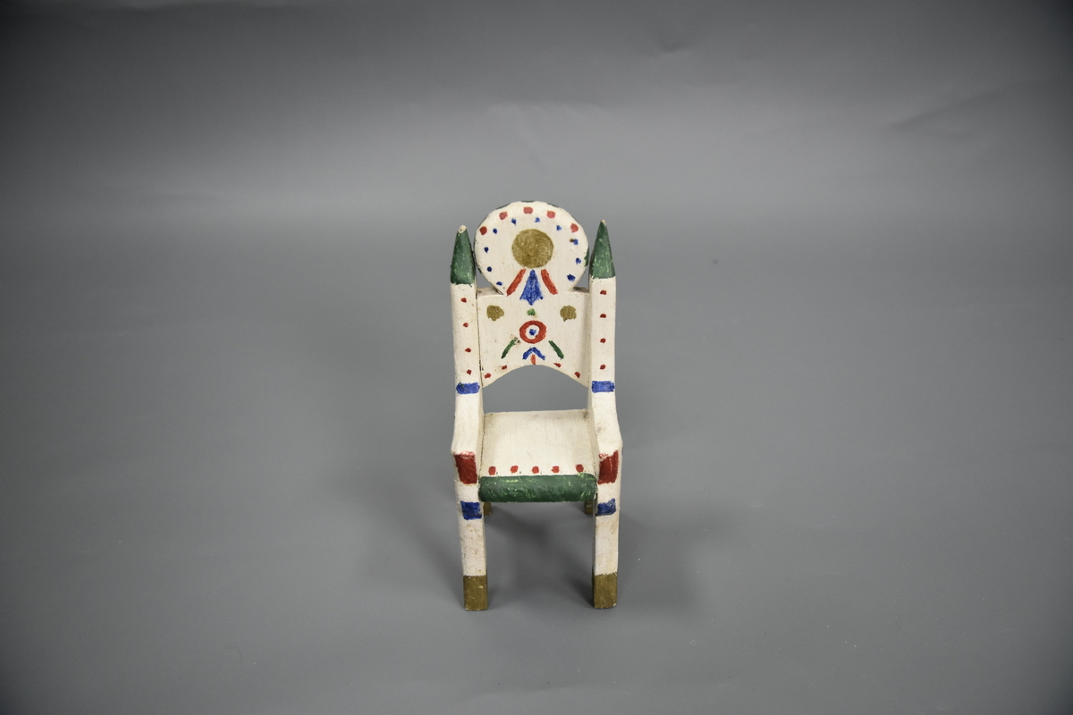 Stol til dokkemøblement i fem deler. Denne stolen er litt mindre enn de tre andre. Møblene er hvitmalte med dekor i ulike farger. 
Møblementet er fra givers tidlige barndom, muligens 1928-1930. Møblene var til hennes første dokkestue, bygget av far av en appelsinkasse. 