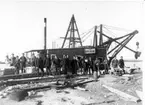 Ett 10-tal män i arbetskläder med skyfflar, skottkärror och stenblock framför ett mudderverk i Gränna hamn. På mudderverket sitter skyltar 