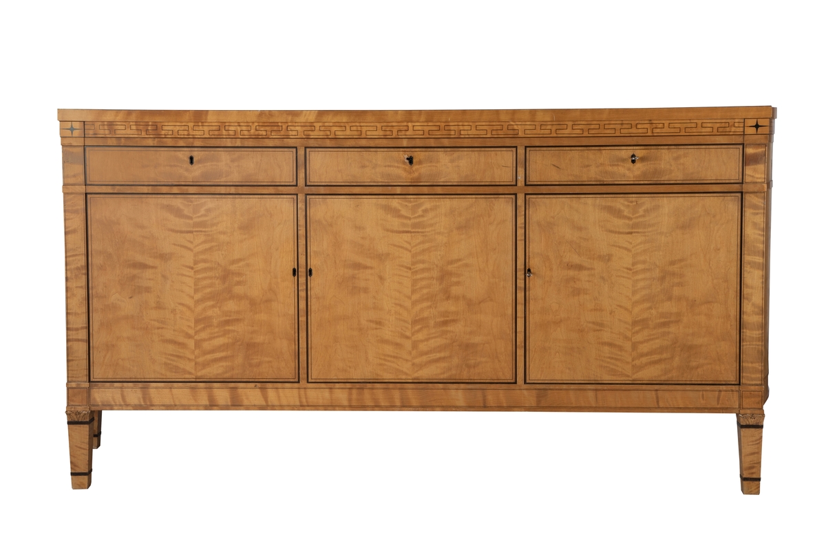 Stor skjenk i lys flammebjørk med intarsia i brunsvart tresort, eventuelt beiset mørk. Rettlinjet, med unntak av endestykkene, som er svakt buet mot en bredere bakvegg. Skjenken har 3 skap og 3 skuffer, hvert skap med 1-2 hyller innvendig, og en smal skuff over. Den midtre skuffen er ødelagt i nøkkelhullet, antagelig etter forsøk på "innbrudd". Nøkler til skuffer og skap. Alle overganger mellom de ulike "etasjer", dvs. mellom ben, understell, skapdører, skuffer, sarg og topplate er markert ved et smalt listverk, som stikker litt utenfor overflaten forøvrig. Topplaten stikker mest ut. Både skuffer, dører, ben og topplate er markert med innlagte smale og bredere konturer og sargen prydet med en gresk, gammel bord, avsluttet med en 4-armet stjerne øverst på "pilastrene" på hver side av fronten. Korte, lett avsmalnende ben, også de med markerte, mørke striper, og utskåret felt med blomst i overgangen mellom ben og skapets understell.