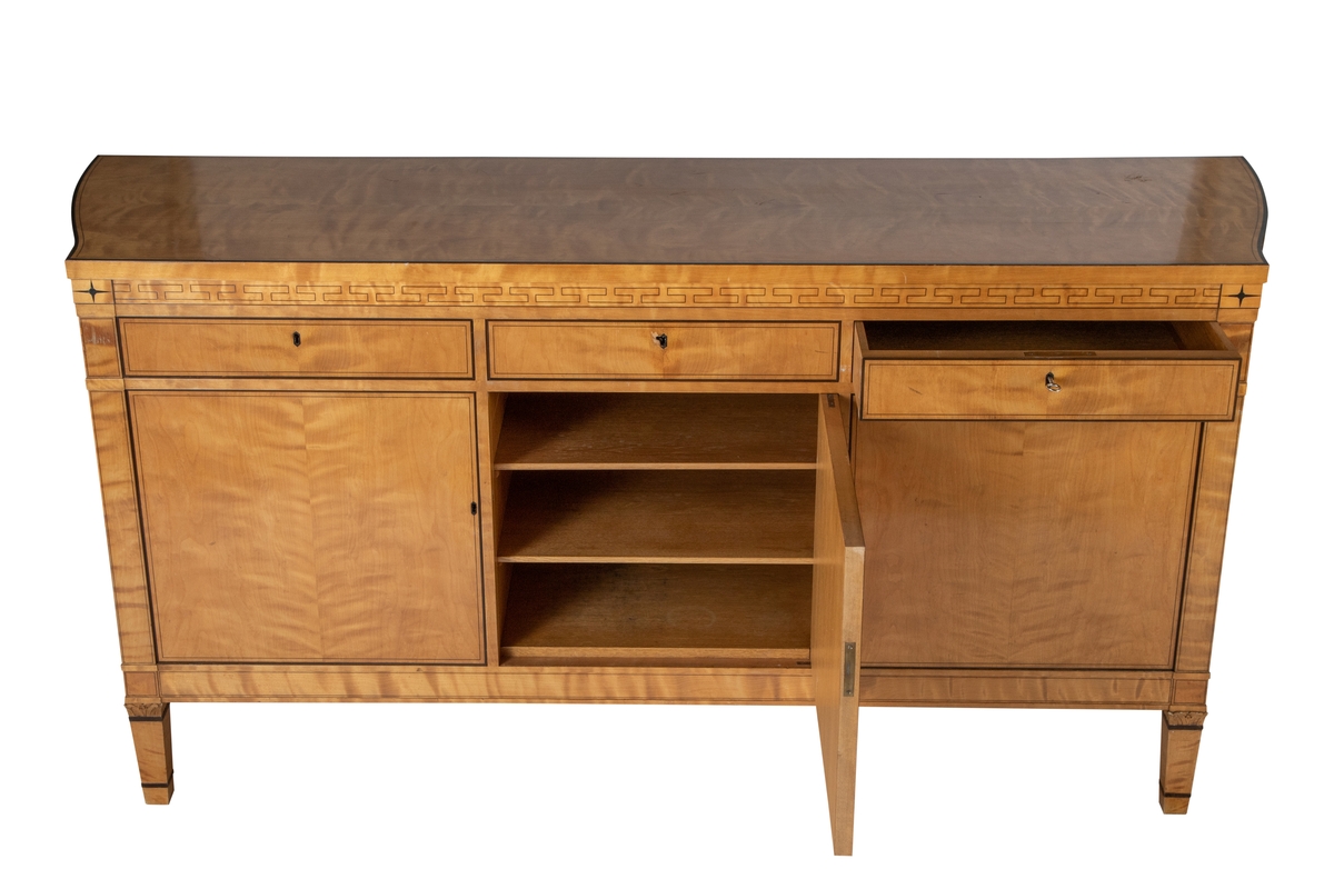 Stor skjenk i lys flammebjørk med intarsia i brunsvart tresort, eventuelt beiset mørk. Rettlinjet, med unntak av endestykkene, som er svakt buet mot en bredere bakvegg. Skjenken har 3 skap og 3 skuffer, hvert skap med 1-2 hyller innvendig, og en smal skuff over. Den midtre skuffen er ødelagt i nøkkelhullet, antagelig etter forsøk på "innbrudd". Nøkler til skuffer og skap. Alle overganger mellom de ulike "etasjer", dvs. mellom ben, understell, skapdører, skuffer, sarg og topplate er markert ved et smalt listverk, som stikker litt utenfor overflaten forøvrig. Topplaten stikker mest ut. Både skuffer, dører, ben og topplate er markert med innlagte smale og bredere konturer og sargen prydet med en gresk, gammel bord, avsluttet med en 4-armet stjerne øverst på "pilastrene" på hver side av fronten. Korte, lett avsmalnende ben, også de med markerte, mørke striper, og utskåret felt med blomst i overgangen mellom ben og skapets understell.