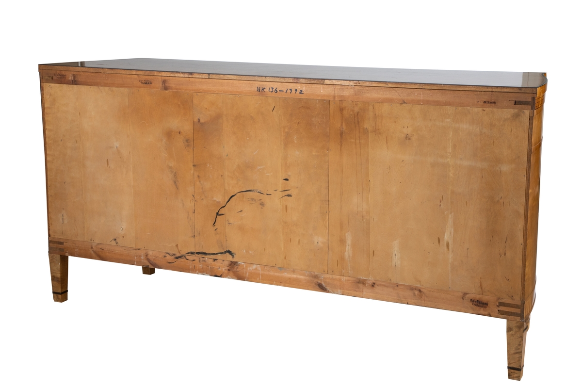 Stor skjenk i lys flammebjørk med intarsia i brunsvart tresort, eventuelt beiset mørk. Rettlinjet, med unntak av endestykkene, som er svakt buet mot en bredere bakvegg. Skjenken har 3 skap og 3 skuffer, hvert skap med 1-2 hyller innvendig, og en smal skuff over. Den midtre skuffen er ødelagt i nøkkelhullet, antagelig etter forsøk på "innbrudd". Nøkler til skuffer og skap. Alle overganger mellom de ulike "etasjer", dvs. mellom ben, understell, skapdører, skuffer, sarg og topplate er markert ved et smalt listverk, som stikker litt utenfor overflaten forøvrig. Topplaten stikker mest ut. Både skuffer, dører, ben og topplate er markert med innlagte smale og bredere konturer og sargen prydet med en gresk, gammel bord, avsluttet med en 4-armet stjerne øverst på "pilastrene" på hver side av fronten. Korte, lett avsmalnende ben, også de med markerte, mørke striper, og utskåret felt med blomst i overgangen mellom ben og skapets understell.