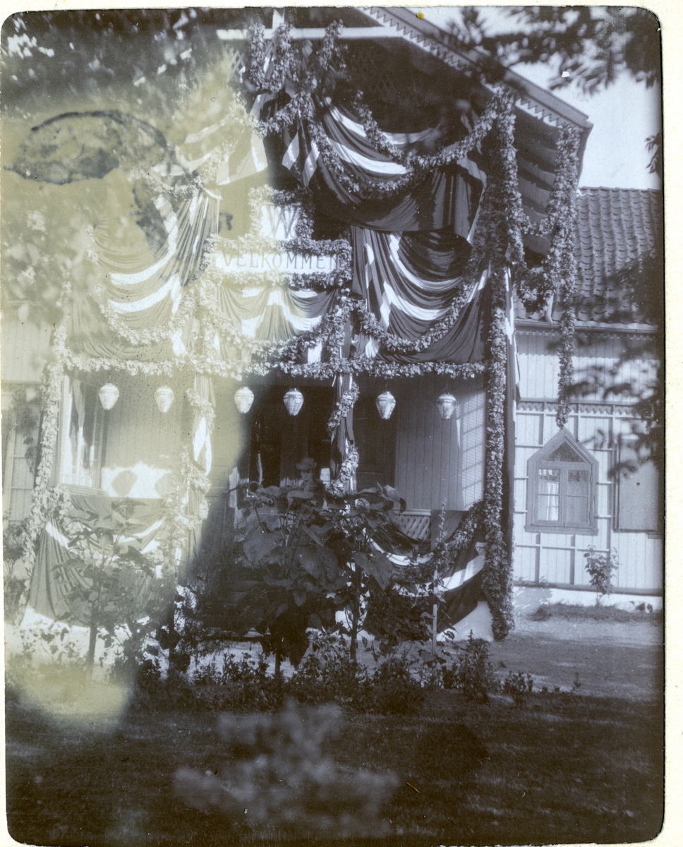 En festpyntet hovedbygning på Egeberg-familiens Strøm gård, til ære for de nygifte Nini og Westye Egeberg. Nini Egeberg skimtes på verandaen. Fotografert september 1904.