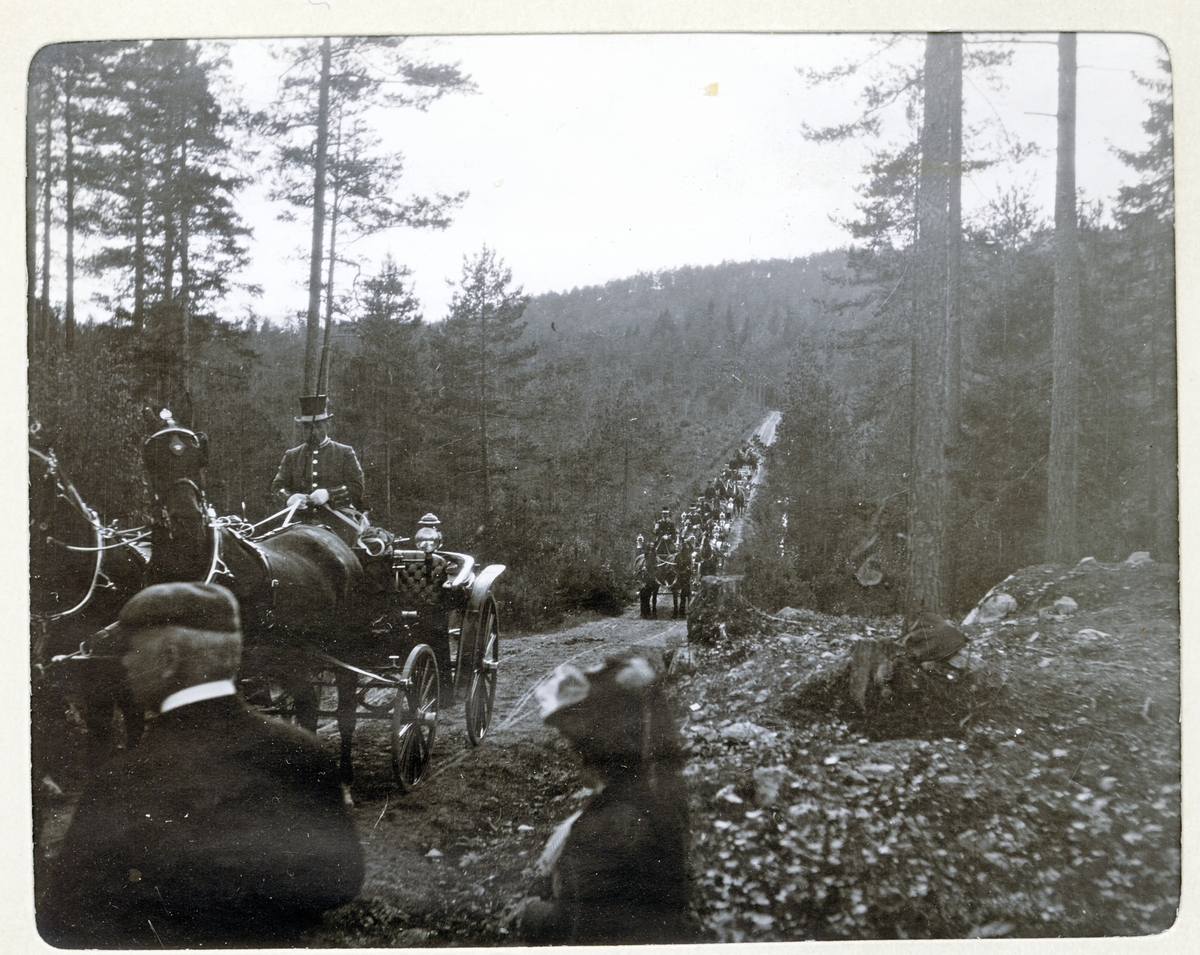 En lang rekke av hestekjøretøyer med kusk frakter elgjegere til eller fra elgjakten på Fossum gård i Bærum(?). Fotografert 1905.