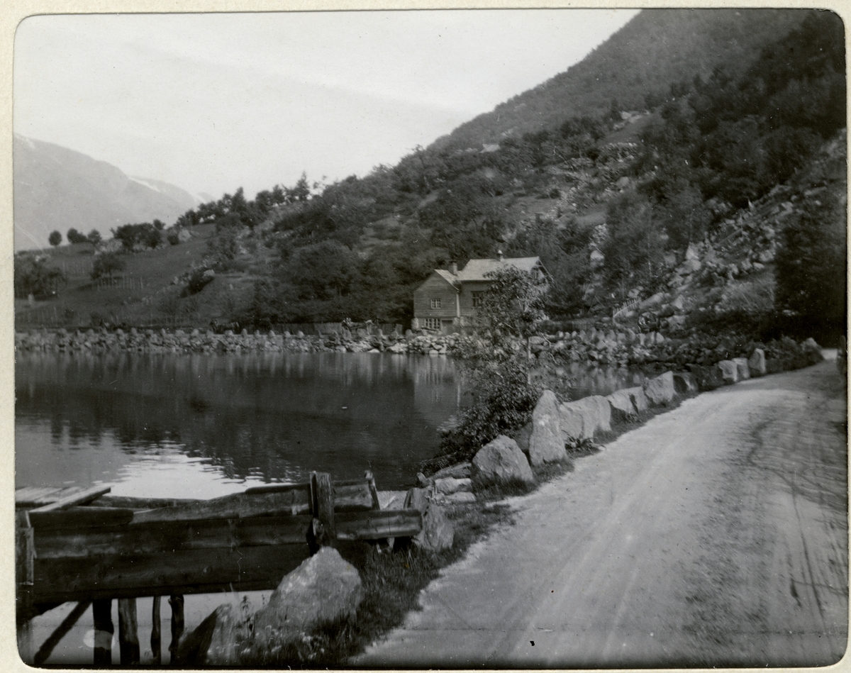 Motiv fra Odda med liten bukt, bolighus, grusvei og rester av en brygge. Fotografert 1906.