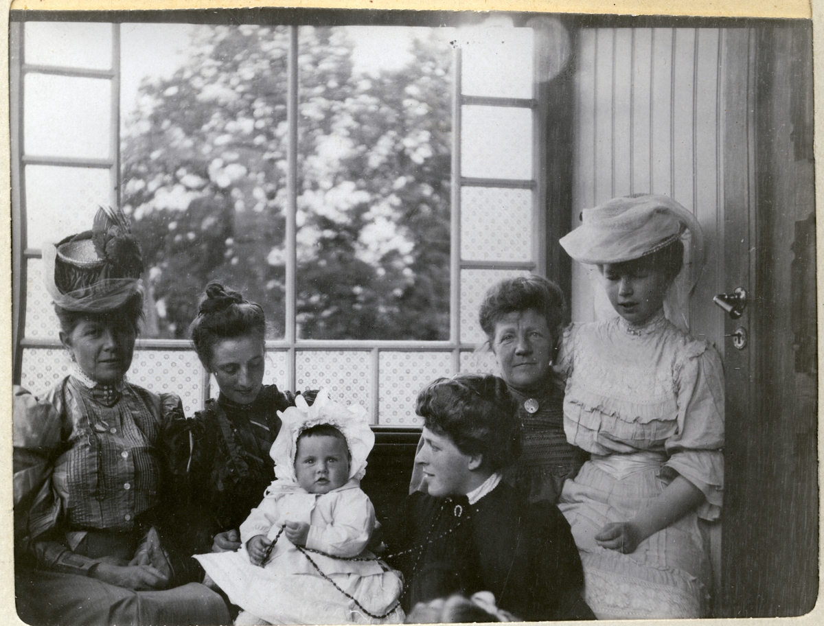 Fem kvinner og liten jente på verandaen til Egeberg-familiens Strøm gård, antagelig Vestre Strøm gård i Rælingen. Nummer to fra venstre er Nini Egeberg med datteren Mimi på fanget. Fotografert august 1906.