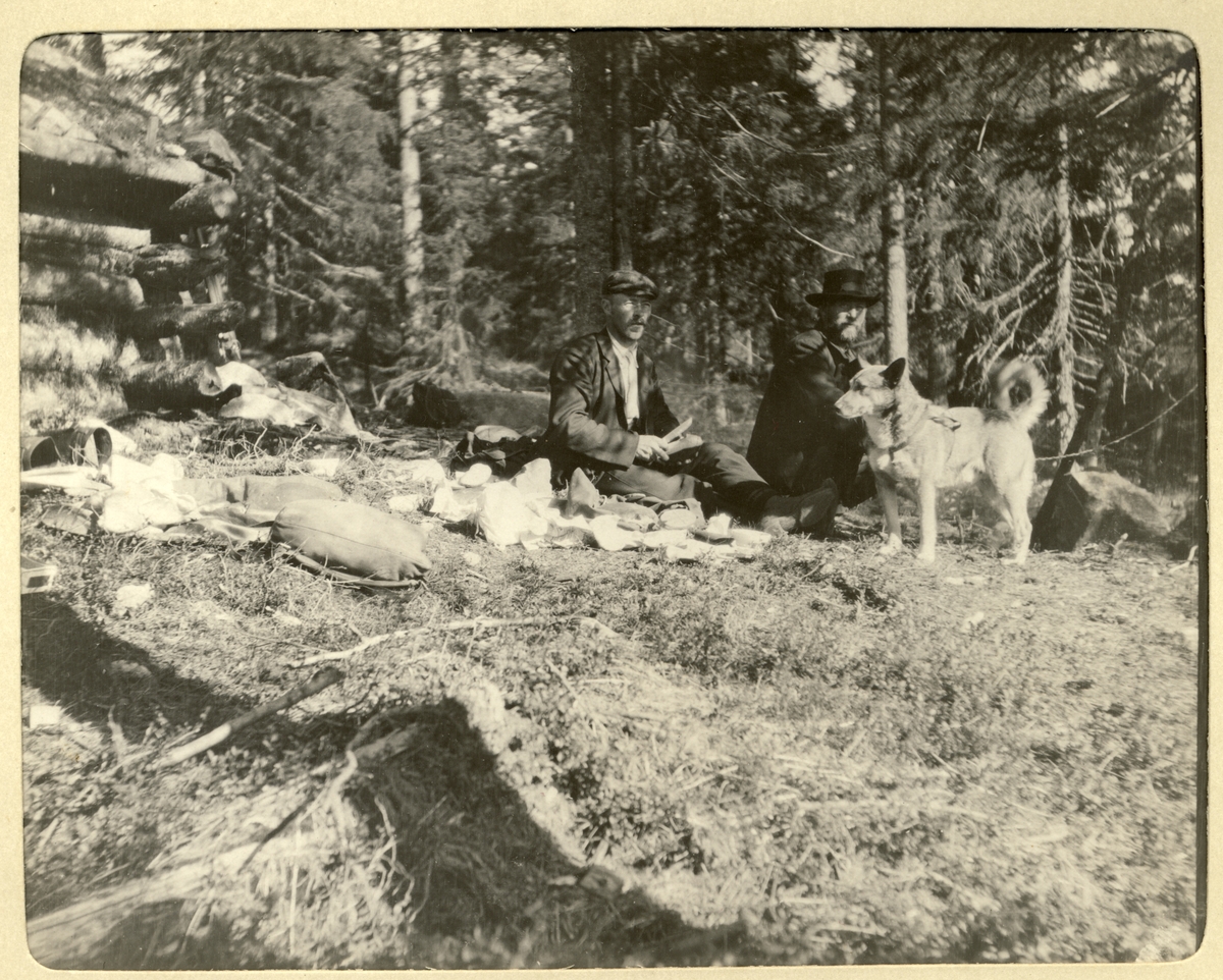 Jegerne Sandbak og Chr. Raaen(?) samt hunden "Russ" tar en spisepause under elgjakten i Romedal, Stange. Til venstre en liten laftet hytte. Fotografert i 1906.