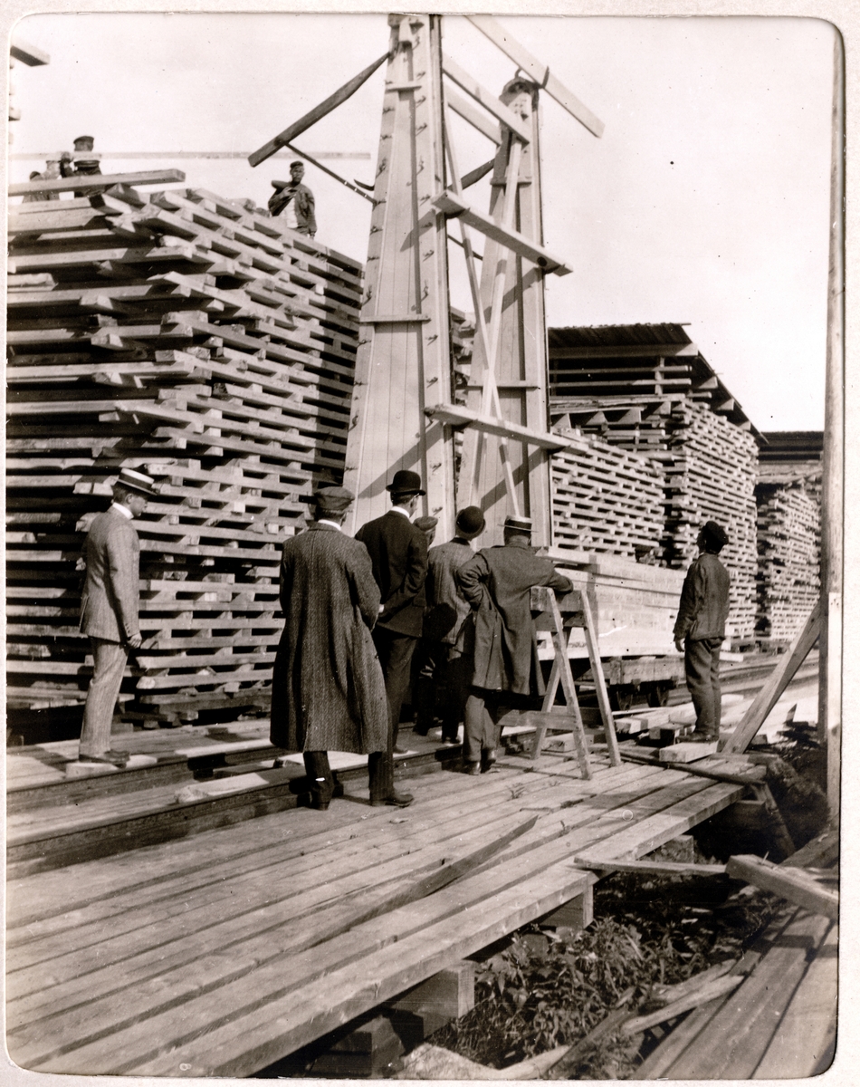 Seks menn inspiserer en plankeløfter på øya Hovinsaara ved Kotka, Finland. På toppen tar to menn imot plankene. Fotografert juli 1907.