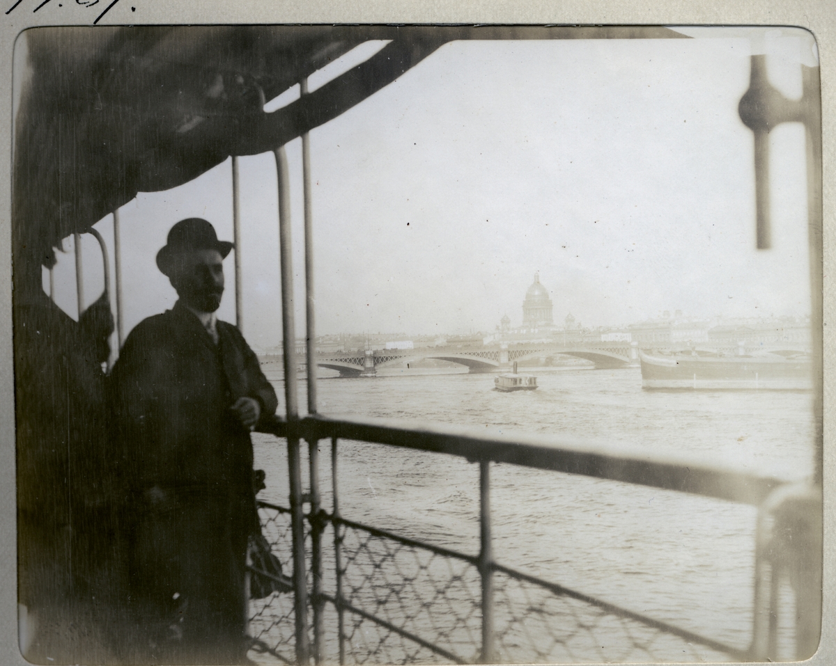 Sekretær Henriksen ved det norske konsulatet i St. Petersburg fotografert på et passasjerbåt på elv i St. Petersburg. I bakgrunnen Anichkov-broen  og Isak-katedralen. Fotografert juli 1907.