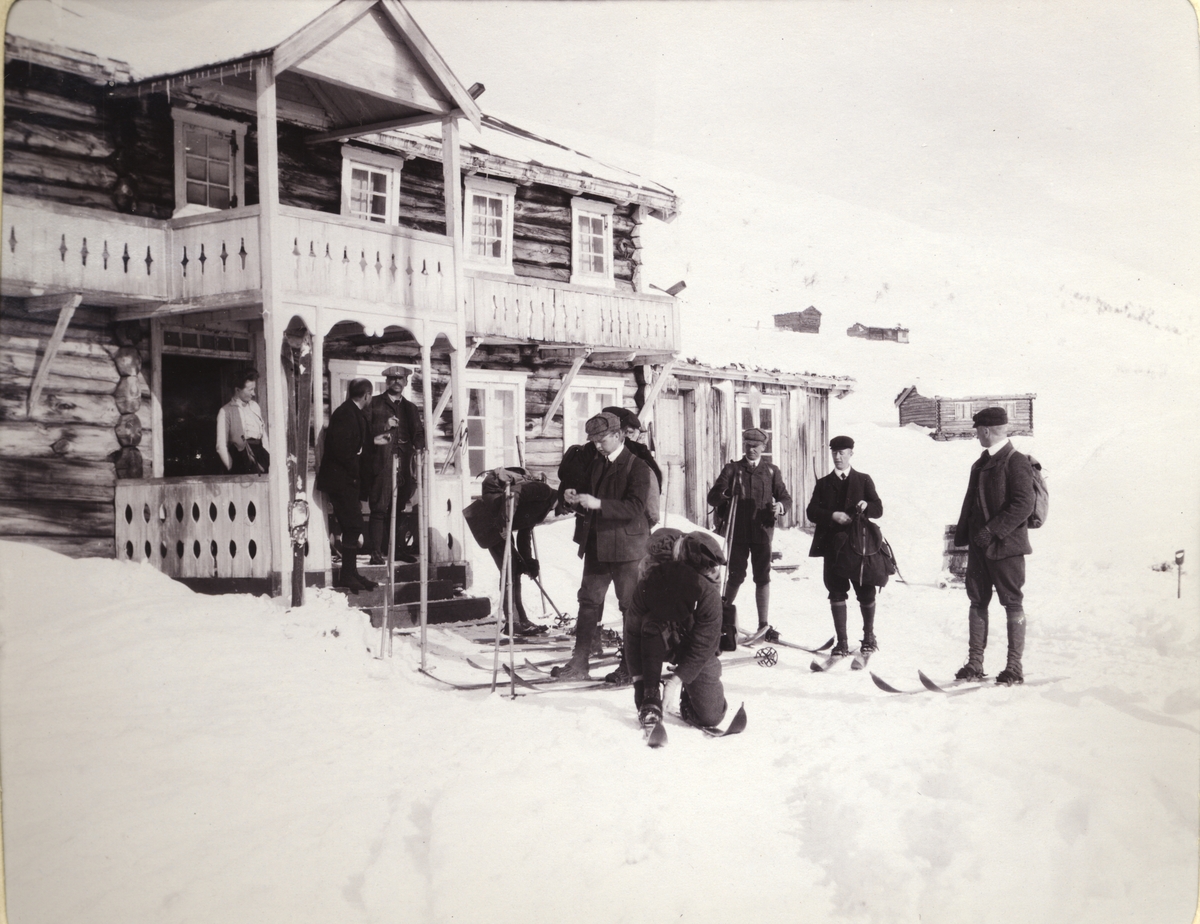 Ni skigåere gjør seg klar til å gå ut fra turisthytten Bessheim i Jotunheimen. En kvinne, kanskje vertinnen, står i døren. Fotografert mars 1908.