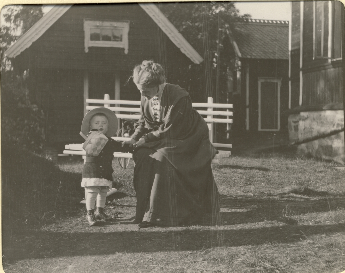 Kvinne, antagelig Ingeborg Fearnley, og et  lite barn, antagelig Thomas Fearnley, på benk ved "Værket"(Mostadværket, Malvik, Nord-Trøndelag?). Fotografert 1909.