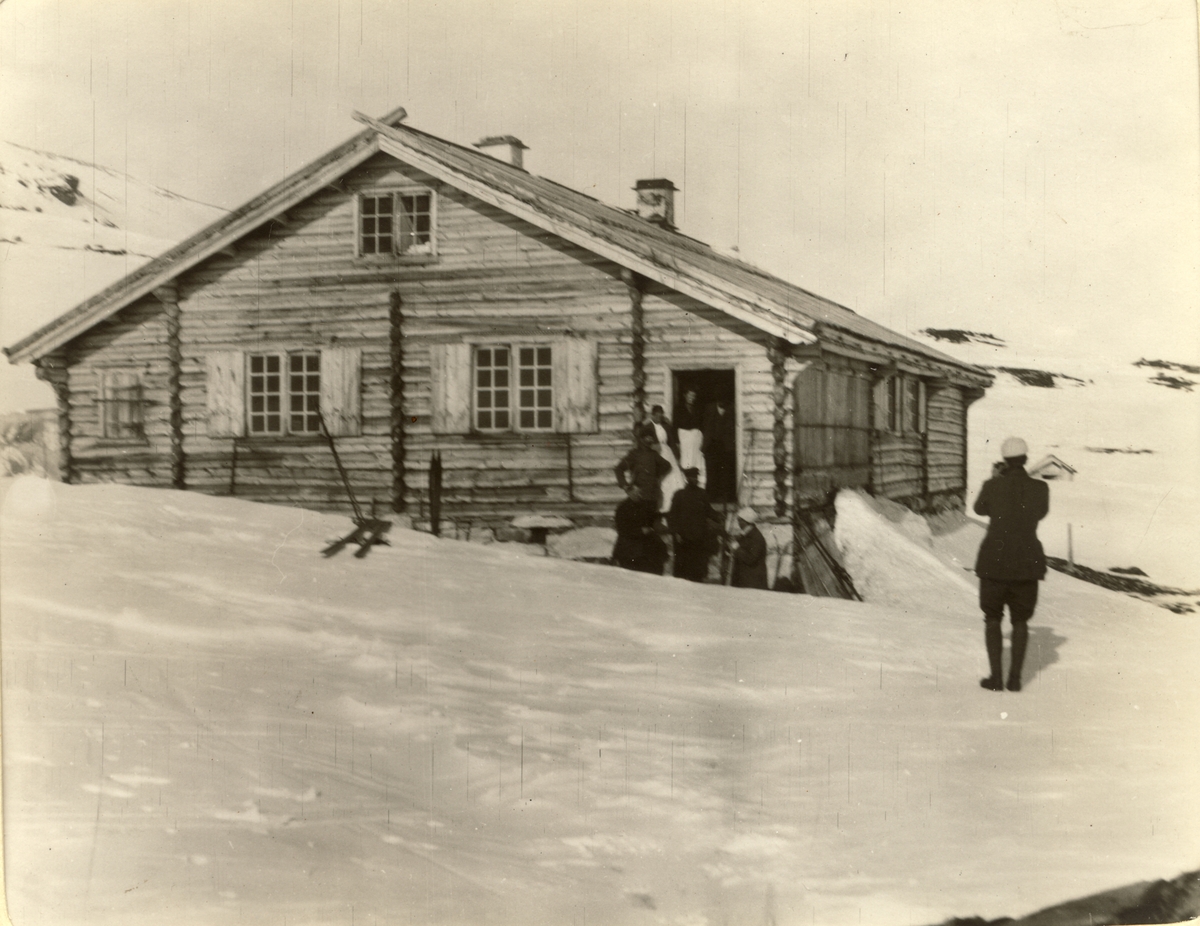 Fem påsketurister blir tatt imot av betjeningen på turisthytten Glitterheim i Jotunheimen. Fotografert påsken 1910.