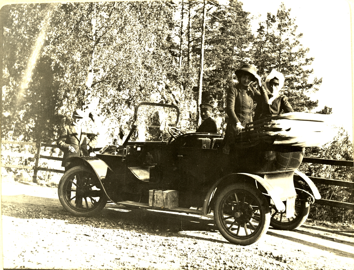 Pause under biltur til Harestua, Søsterkirkene, Hønefoss. Her ved "Skaret" i Hole, Buskerud. I bilen står Cessy von Wedel og Nini Egeberg. Carl Løvenskiold(?) står foran bilen. I bilen sitter dessuten sjåføren. Fotografert juli 1911.