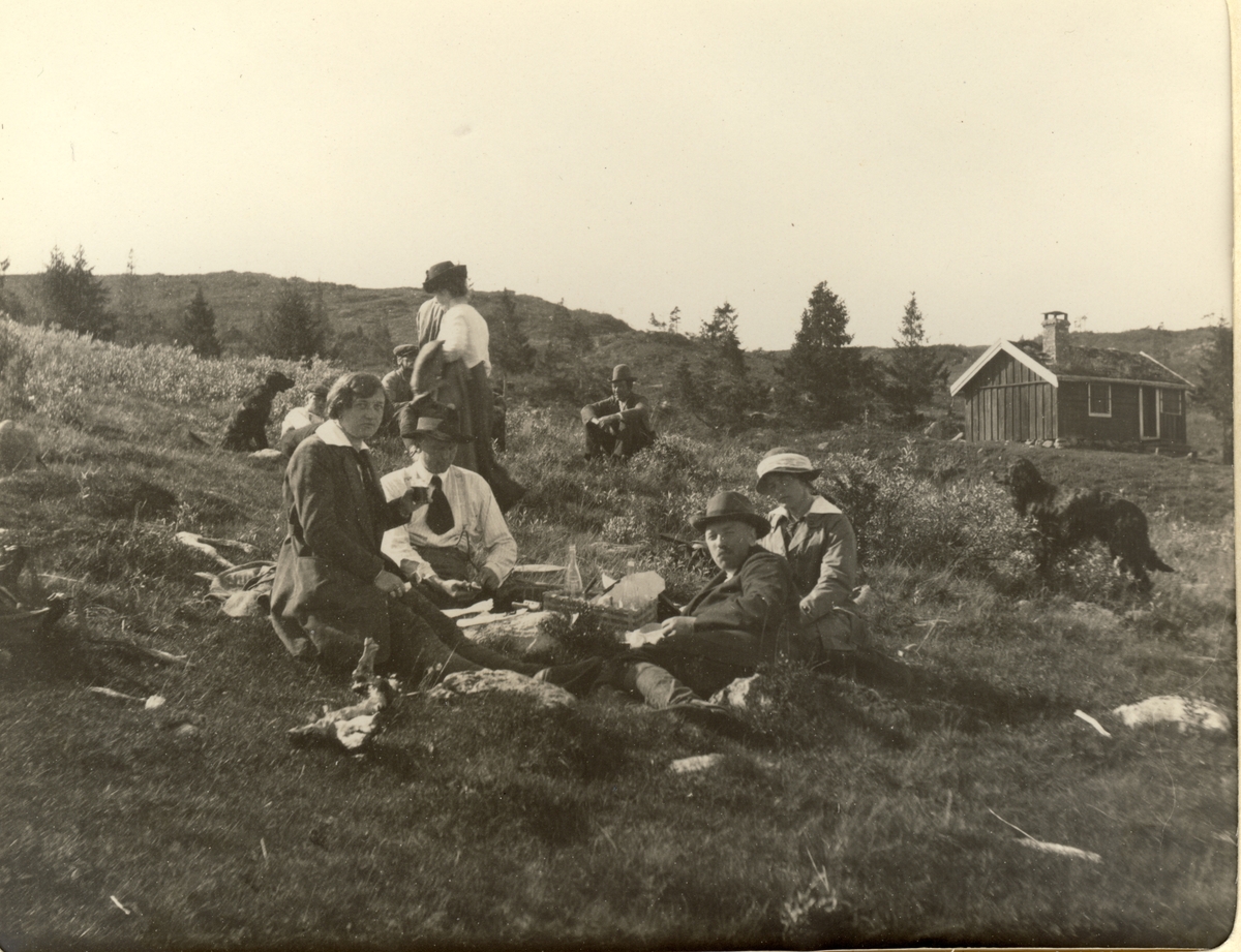 Gjester, medhjelpere og jakthunder fra Fearnley-familiens jakthytte ved Øst-Fjergen raster underveis. Liten hytte i bakgrunnen. Fotografert 1913.