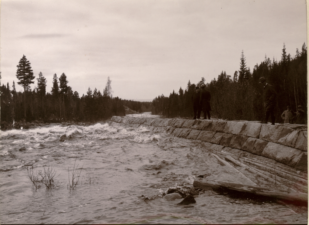 Tømmerfløting antagelig i Osfallet, Åmot, Hedmark. Medlemmer av Christiania Tømmerdirektion står på elvebredden. Fotografert i forbindelse med befaring i mai 1915.