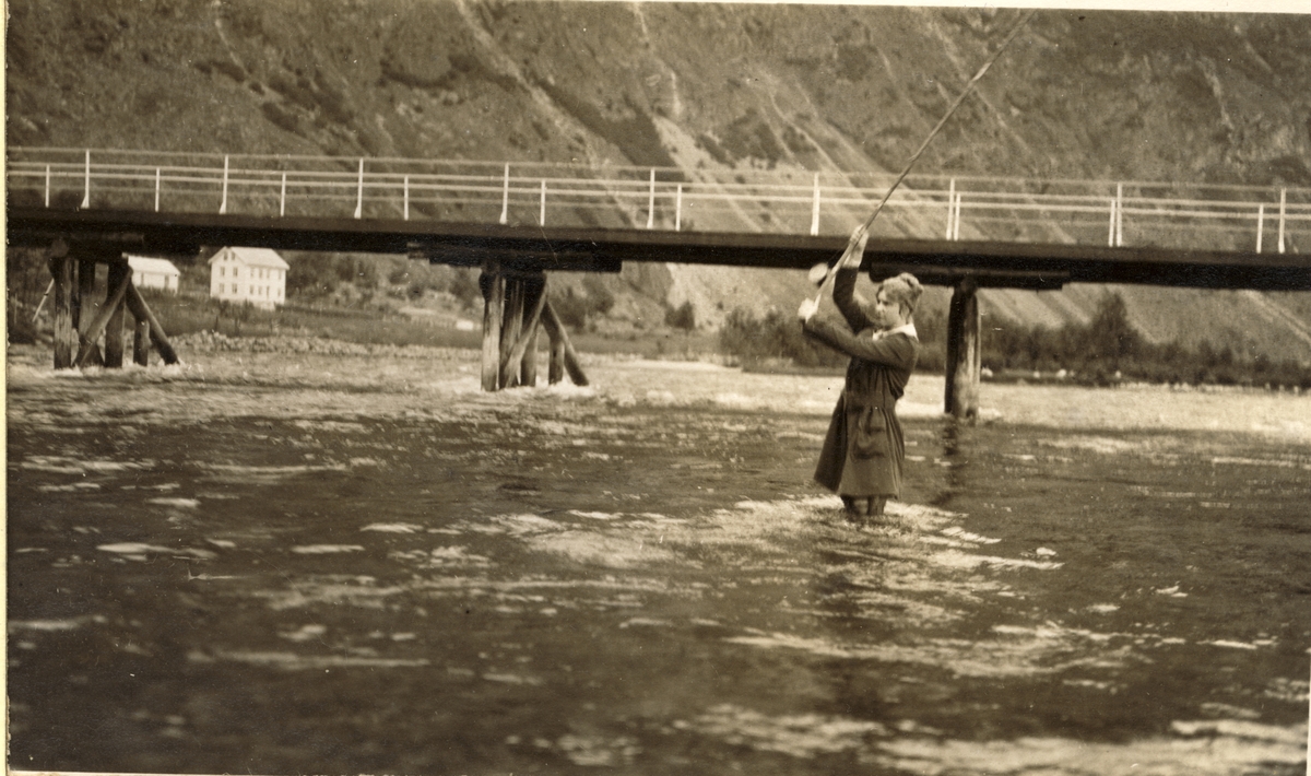 Evelyn Egeberg fisker i Lærdalselvi, Sogn og Fjordane. I bakgrunnen en bro over elven og et gårdstun. Fotografert sommeren 1916.