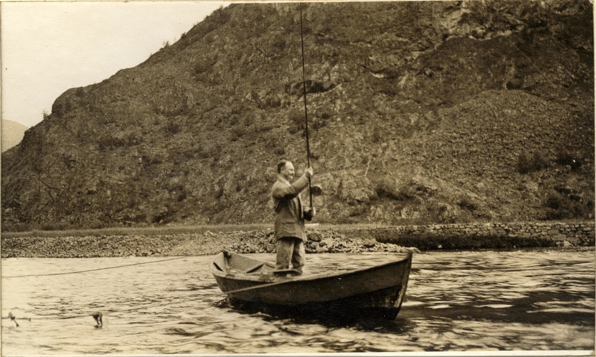 Waldemar Kallevig, stående i robåt, har antagelig fått en laks på kroken i Lærdalselvi, Sogn og Fjordane. I bakgrunnen går en vei langs med fjellet. Fotografert sommeren 1916.