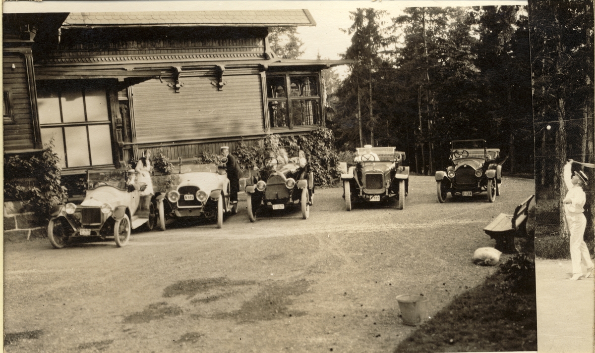 Fem biler er parkert foran trevilla, antagelig Ferdinand J. Egebergs villa på Snarøya. I noen av bilene sitter det sjåfører og passasjerer. Fotografert 1916.