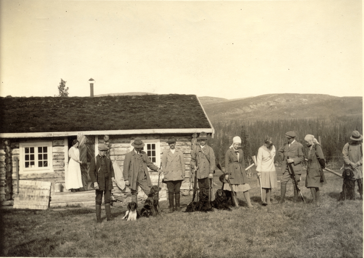 Jaktlag og medhjelpere samlet foran Fearnley-familiens jakthytte ved Øst-Fjergen i Meråker. En kvinnelig medhjelper står ved døren i bakgrunnen. Fotografert 1916.