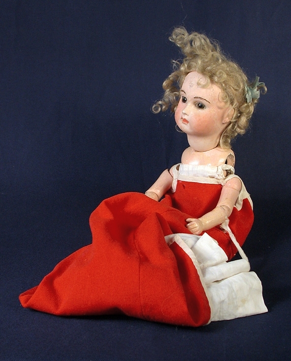 :1 Docka, massivt huvud av gips, övriga kroppsdelar av papier machè. Ledkulor av trä. Ögon av glas. Blont lockigt hår. 
:2 Klänning, i röd tuskaftsvävd ylle. 
:3 Underklänning lång, vid av linne. 
:4-5 Skor