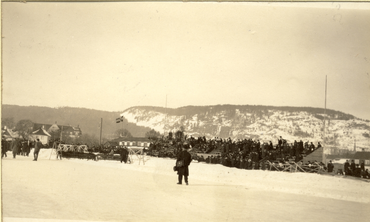 Banen på Marienlyst i Drammen blir benyttet til ridestevne. Tilskuere sitter klar på tribunen og flagget er heist. I bakgrunnen bolighus og hindre til sprangridning. Fotografert mars 1917.