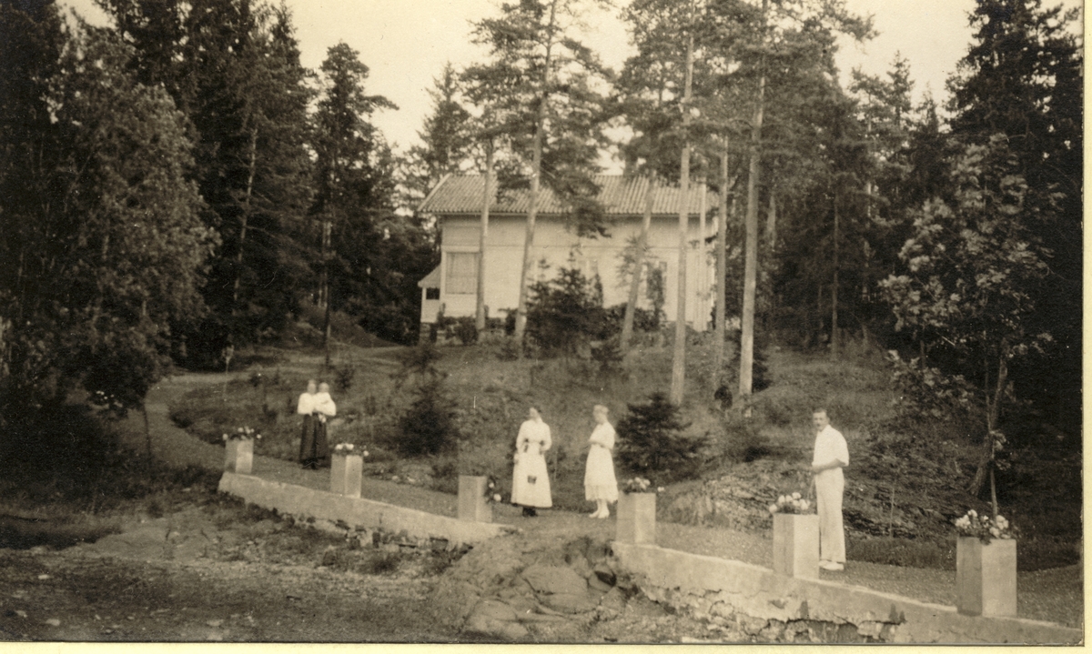 Tre kvinner, én med barn på armen, og en mann foran Severin Finnes villa på Grimsøya i Bærum. Mannen til høyre er antagelig Severin finne og kvinnen med barn antagelig hans kone Esther, f. Egeberg, med sønnen Hans-Gabriel. Fotografert 1917.