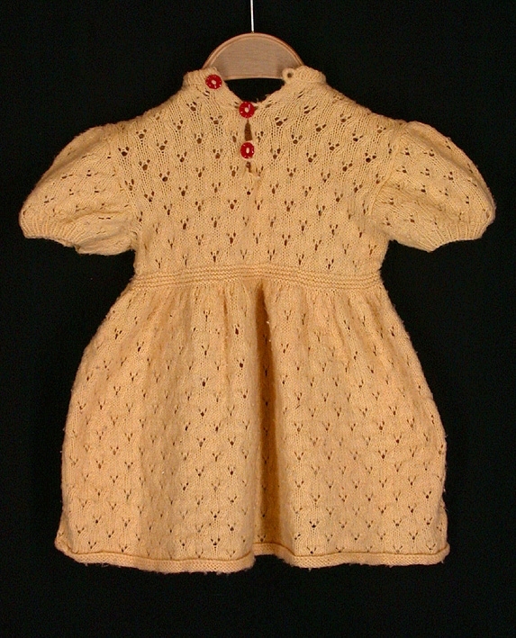 Gul, hemstickad barnklänning. Slätstickad i hålmönster med resårstickning i ärmslut, halsringning, midja och kjolens nederkant. Kort ärm med liten puff. Sprund i halsen fram med 3 st röda plastknappar och stickade hällor.