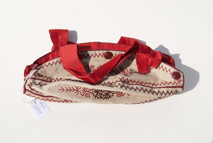 Liten väska för stickning. Bottentyg i beige linnetyg. 5 st ovala tygbitar hopsydda med passpoalerad rött linne mellan bitarna samt runt ytterkanten. Broderad med mönster, blommor, snirklar i rött och brunt med stjälkstygn och kråkspark. 3 st bruna knappar på ena långsidan andra en virkad trådögla övriga saknas. Fodrad med rött linnetyg. Handtag av rött sidenband avslutad med rosett.
Användes för att bära med sig liten stickning.