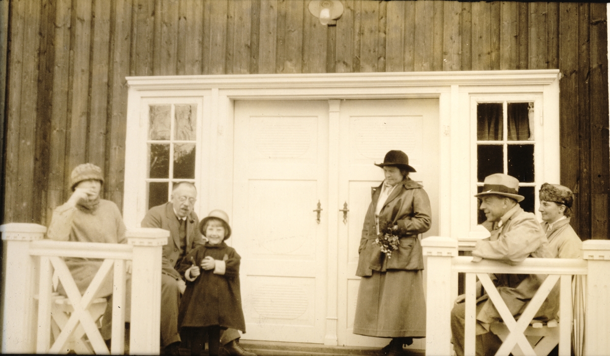 Fem voksne og et barn på verandaen til Brekke gård(?). Helt til høyre sitter Nini Egeberg. Fotografert pinsen 1923.