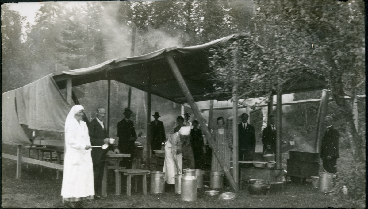 Et provisorisk kjøkken satt opp på gårdsplassen til jaktslottet Malungen, i forbindelse med en folkefest 22. juni 1924. Under teltduken står kvinnelige og mannlige medhjelpere. En kvinnelig kokk til venstre med suppeøse.