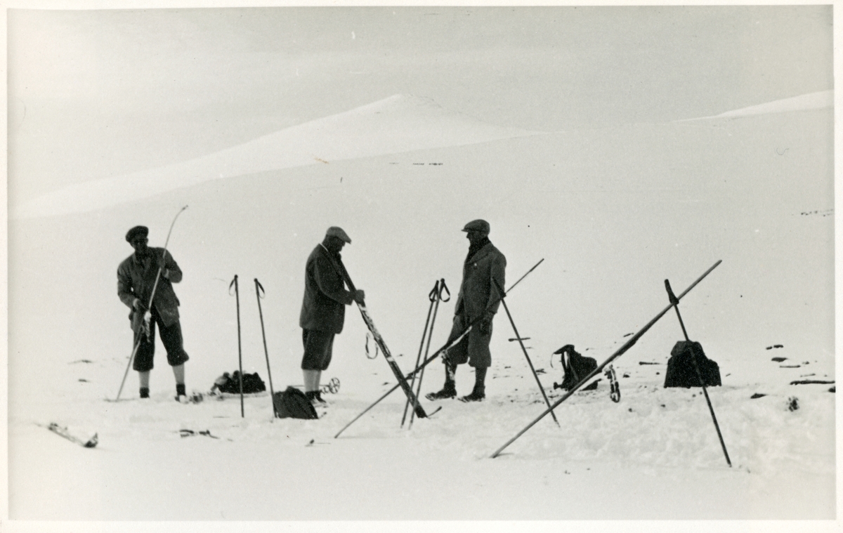 Tre skigåere holder pause for å smøre ski på vei over Russlirundhøe. Fra venstre "Bass", Westye Egeberg og Søren Sommerfelt.  I bakgrunnen ses Nautgardstind. Fotografert 1934.