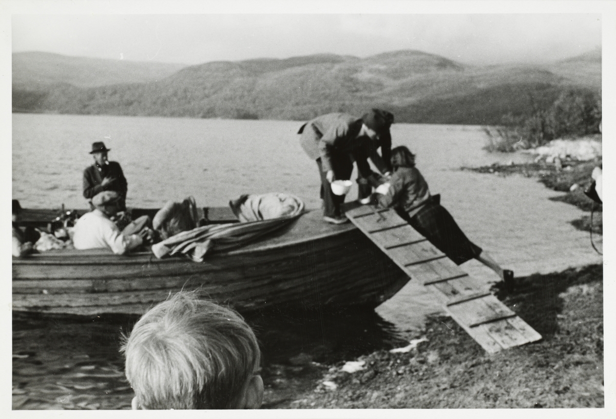 En kvinne og en mann hjelper en hund om bord i en trebåt. Andre passasjerer venter i båten og på land. Antagelig ved Møsvatn i Vinje, Telemark.