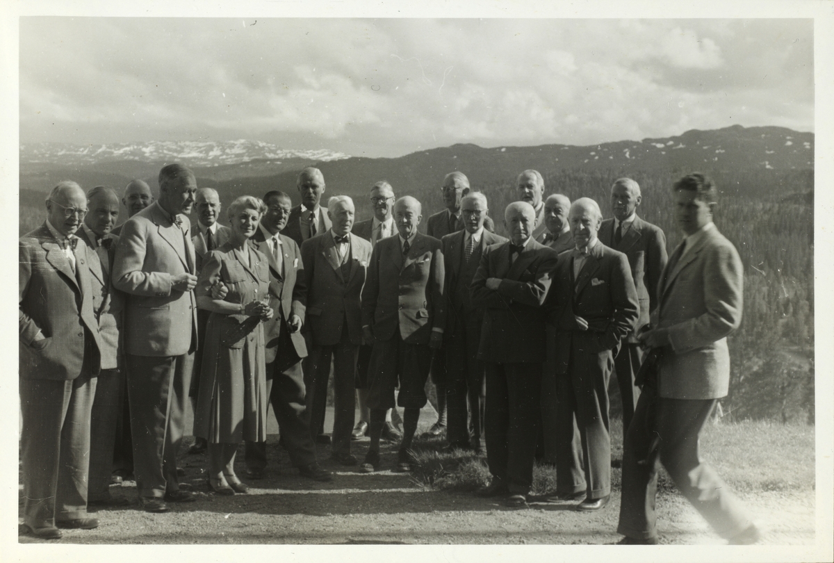 Gruppebilde av medlemmer av skiklubben Fram på besøk hos direktør for Orkla Thorry Kiær og hans kone Ingrid i Trøndelag. Her ved Fjeldheim gård i Meldal. Fjellandskap i bakgrunnen. Fotografert 1952.