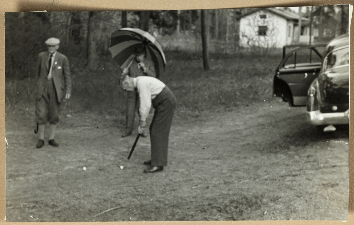 Tre menn fordriver tiden med noen slag golf mens de venter på flere deltakere. Til venstre Aksel Hansen, i midten August Walle Hansen med parasoll/paraply, og foran Thorry Kiær med golfkølle.  Antagelig fotografert i nærheten av Dejefors, Sverige, mens skiklubben Fram var på besøk der i mai 1954.