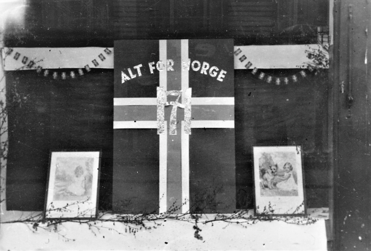 Feiring av freden 1945. Vindusutstilling. I midten av bildet henger et Norsk flagg med "ALT FOR NORGE" øverst og "Håkon 7. logen" over krysset i senter av flagget. På hver side et telegram. Små norske flagg i bue tverrs over vinduet.