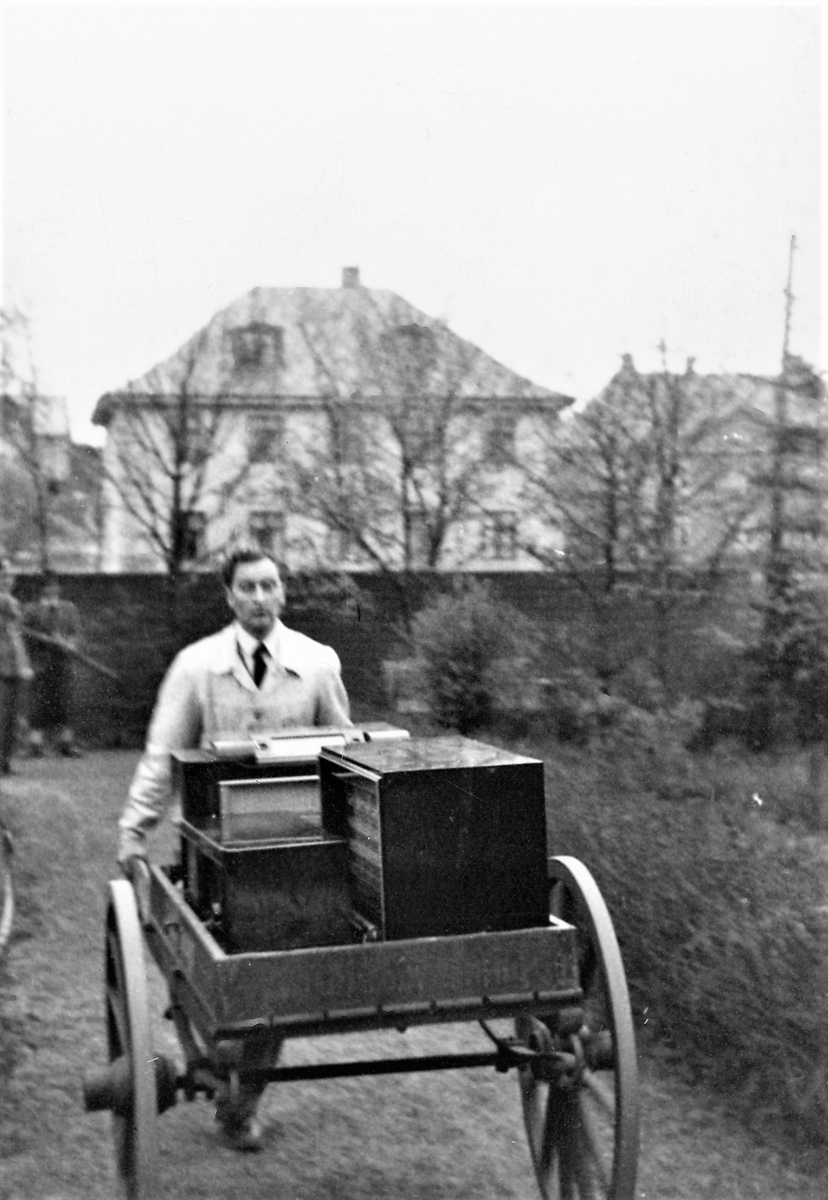 Feiring av freden 1945. Mann med håndkjerre som frakter radioer. To motstandsfolk til venstre i bakgrunnen. Trær og en mur foran en bygningsrekke. Trolig fra hjemhentingen av lagrede radioapparater i Festiviteten den 8. mai