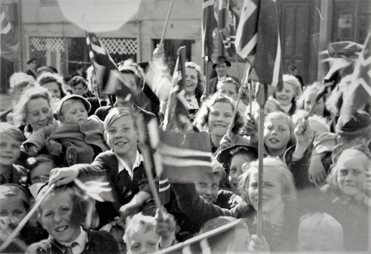 Feiring av freden 1945. En større forsamling av festkledde barn med norske flagg feirer freden og trolig et kongelig besøk. Det ser ut som barna står langs en traser der de skal få se enten kronprins Olav kjøre forbi, eller Haakon den VII.